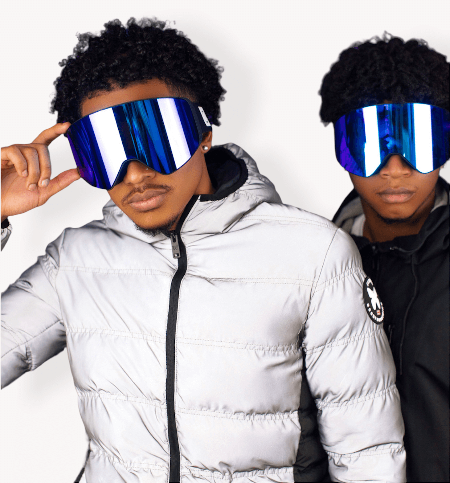 Two men posing with puffer coats: one a reflective silver, the other black. Both are wearing the Icy Blue CJ Goggles