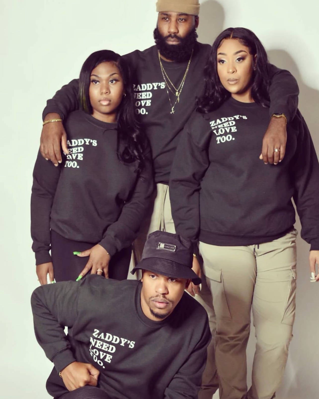 A group photo of two women and two men. All are wearing black crew neck sweaters that say, "Zaddy's Need Love Too." One man has on a Christian James Bucket Hat in black.