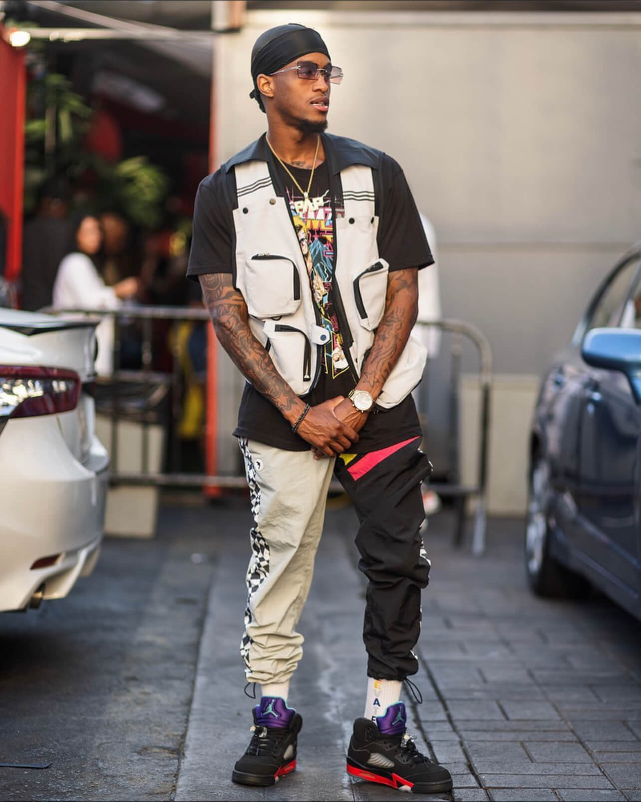 A man posing with a black durag, graphic tee and vest, adoring a pair of CJ frames. He's also wearing a popular timepiece of the brand called Varsity.
