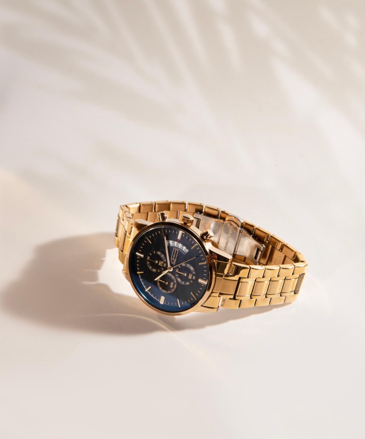 An up-close photo of the timepiece Trident. The watch has a round blue face and a striking gold link band. The hands, hour markers, and dials are also gold. 